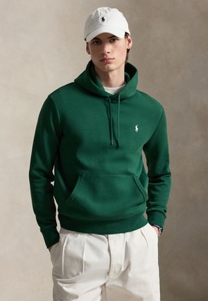Junger Mann trägt einen dunkelgrünen Hoodie mit Fronttasche und weißem Logo, weiße plissierte Hose und eine weiße Baseballkappe vor einfarbigem Hintergrund.