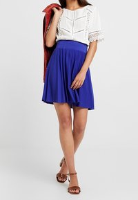 White, short-sleeve blouse with lace accents and buttons paired with a pleated, knee-length, royal blue skirt. Brown heeled sandals.