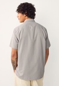 Homme aux cheveux bouclés portant une chemise à manches courtes grise claire à motifs et un pantalon beige, de dos avec les mains dans les poches, tatouages visibles sur les bras.