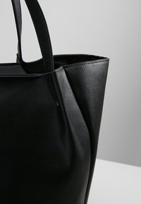 Sac fourre-tout en cuir noir avec deux anses, montré en gros plan sur un fond gris neutre.