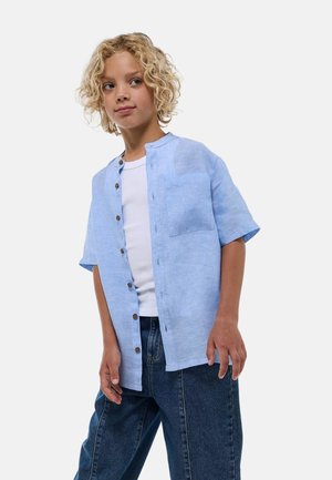 Chemise bleu clair à manches courtes avec boutons, col rond, une poche poitrine et tissu texturé. Portée avec un jean en denim bleu.