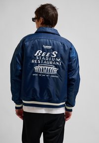 Navy satin bomber jacket featuring white embroidered text and graphic on the back. Ribbed cuffs and hem, with contrasting stripes.