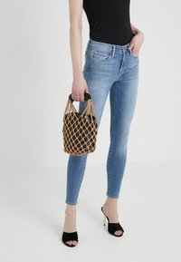 Czarna pleciona torba z brązowymi sznurkami, noszona z jasnoniebieskimi dopasowanymi dżinsami i czarnymi sandałami na obcasie, ukazująca elegancki, nowoczesny design.