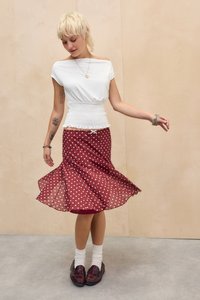 Tânără cu păr blond scurt care se învârte, purtând un top alb cu smocuri, fustă roșie cu buline, șosete albe și mocasini burgundy.