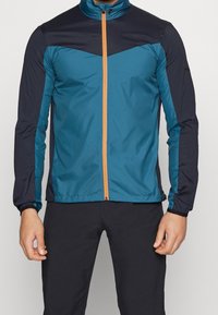 Vindjakke med en teal krop, marineblå skuldre og en orange frontlynlås. Letvægtigt, struktureret stof med elastiske ærmer.