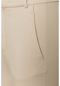 Gros plan d’un pantalon beige montrant une poche latérale, une passants de ceinture et une texture de tissu lisse.