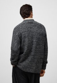 Teksturowany szary wełniany sweter o luźnym kroju, z szerokimi rękawami i czarną metką na karku. Delikatny wzór na całej tkaninie.