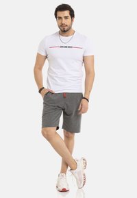 Weißes T-Shirt mit schwarzem und rotem Text, graue Shorts mit verstellbarem Bund und weiße Turnschuhe mit roten Akzenten.