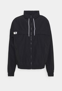 Sort vindjakke med høj krave, hvide snøre, lynlås foran, elastiske manchetter og en kontrastfarvet logo-patch på brystet.
