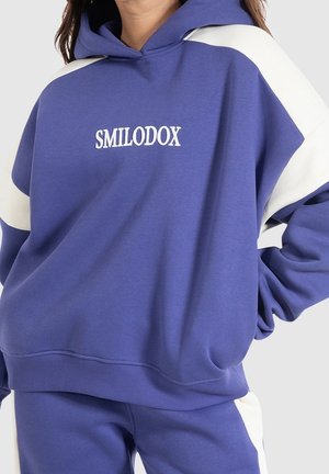 Felpa con cappuccio viola con inserti bianchi sulle spalle e la scritta "SMILODOX" ricamata in bianco. Tessuto in misto cotone con vestibilità comoda.