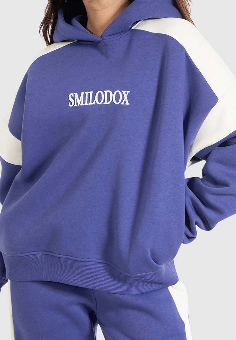 Purple hooded sweatshirt featuring white shoulder accents and "SMILODOX" embroidered in white. Cotton blend fabric with a relaxed fit.