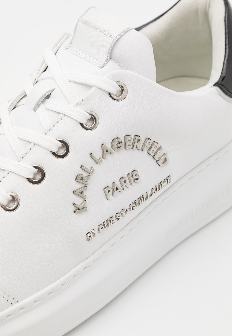 Sneaker en cuir blanc avec un logo argenté, œillets en métal ronds et lacets blancs. Présente un capuchon de talon noir texturé et un col rembourré.