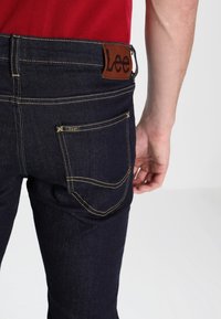 Ciemnoniebieskie dżinsy z żółtym przeszyciem, brązową skórzaną łatą z logo „Lee” oraz tylną kieszenią z minimalnym zdobieniem.