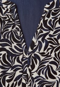 Bluse mit V-Ausschnitt, die einen marineblauen Hintergrund mit schwarzen und blauen Blattmustern aufweist. Der Stoff hat eine strukturierte Optik mit sichtbaren Falten.