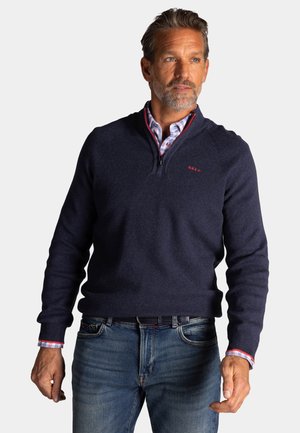 Man draagt een marineblauwe trui met een kwart ritssluiting over een geruit overhemd en een blauwe spijkerbroek, kijkt iets naar de zijkant tegen een effen achtergrond.