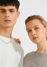 Nuori mies ja nainen, jotka ovat pukeutuneet kultaketjuihin, seisovat lähellä toisiaan neutraalit ilmeet kasvoillaan. Naisen käsi lepäilee miehen olalla.