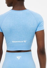 Zadní pohled na osobu v jasně modrém sportovním krátkém topu a legínách s vysokým pasem se bílým nápisem "Squatproof".