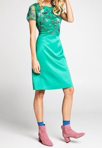 Zelený smaragdový overal s průhledným, vyšívaným topem se květinovými vzory. V kombinaci s růžovými kotníkovými botami a modrými ponožkami.