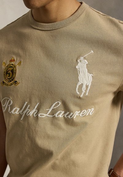 Μπεζ πόλο Ralph Lauren με λευκό κεντημένο λογότυπο παίκτη πόλο πάνω σε άλογο και έμβλημα ασπίδας στο στήθος.