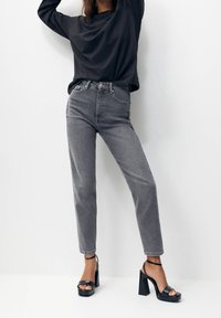 Jeans grigi alti in vita realizzati in denim, abbinati a una maglietta nera a maniche lunghe e sandali neri con plateau. I jeans presentano un classico design a cinque tasche.