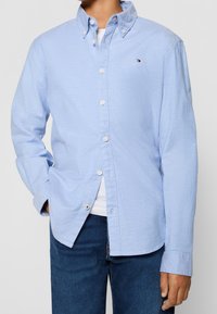 Mann in einem hellblauen, knöpfen Hemd mit langen Ärmeln über einem weißen T-Shirt und dunkelblauen Jeans, Hand in der Tasche, vor einem schlichten Hintergrund.