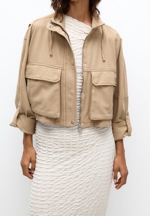 Femme portant une veste courte beige avec de grandes poches avant sur une robe blanche texturée longueur genoux.