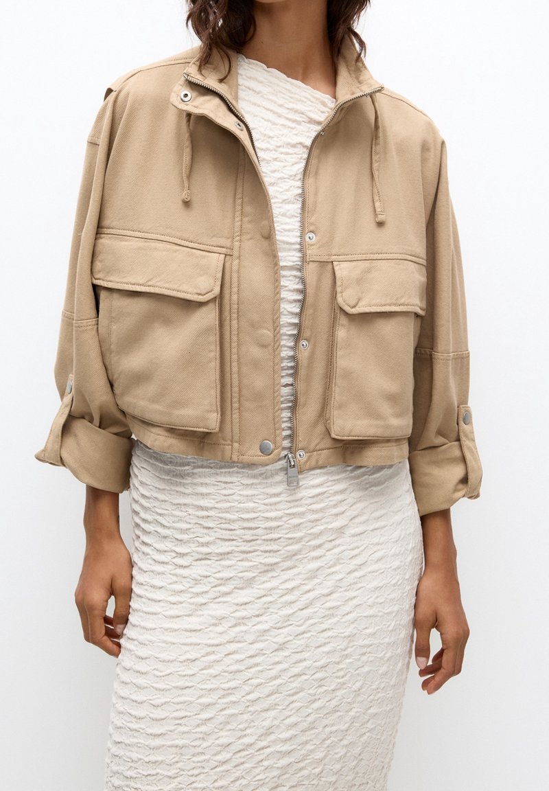 Femme portant une veste courte beige avec de grandes poches avant sur une robe blanche texturée longueur genoux.