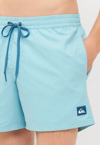 Pantaloni scurți de înot de culoare albastru deschis, confecționați dintr-un material neted, având șnur pentru ajustare și buzunare laterale. Include un logo al mărcii la margine.