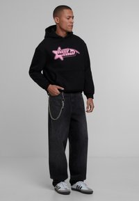 Sudadera negra con gráfico rosa que presenta una estrella, combinada con jeans oscuros holgados y zapatillas blancas con franjas negras. Detalle de cadena plateada visible.