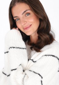 Weißer, gerippter Strickpullover mit schwarzen Streifen, der lockere Ärmel und eine weiche Textur hat. Das Modell hält den Pullover sanft.