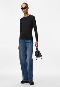 Sort langærmet skjorte, lyseblå vide bukser, sorte solbriller, holder en lille mørk taske med snøring.