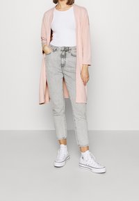 Cardigan largo rosa claro, top blanco, jeans de cintura alta gris y zapatillas blancas, de pie sobre un fondo gris.