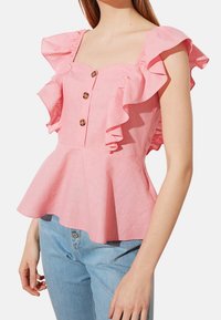 Top peplum rosa con maniche a balze, scollatura quadrata e tre bottoni marroni sul davanti. Indossato sopra jeans blu chiaro.
