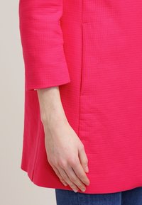 Kirkkaan vaaleanpunainen, tekstuurilla varustettu tunika, jossa on kolme neljäsosaa hihat, rento istuvuus ja näkyvät sauman yksityiskohdat. Käsivarsi nojaa vaatteeseen.