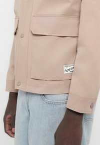 Μπεζ μπουφάν με τσέπες με καπάκι στο μπροστινό μέρος και μεταλλικά κουμπιά πατιλέτας, φορεμένο πάνω από ανοιχτόχρωμο μπλε τζιν σε άτομο με σκούρο δέρμα.