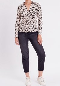 Mujer vistiendo una camisa de manga larga con frunces y estampado, jeans cortos negros y zapatillas blancas, de pie contra un fondo liso.