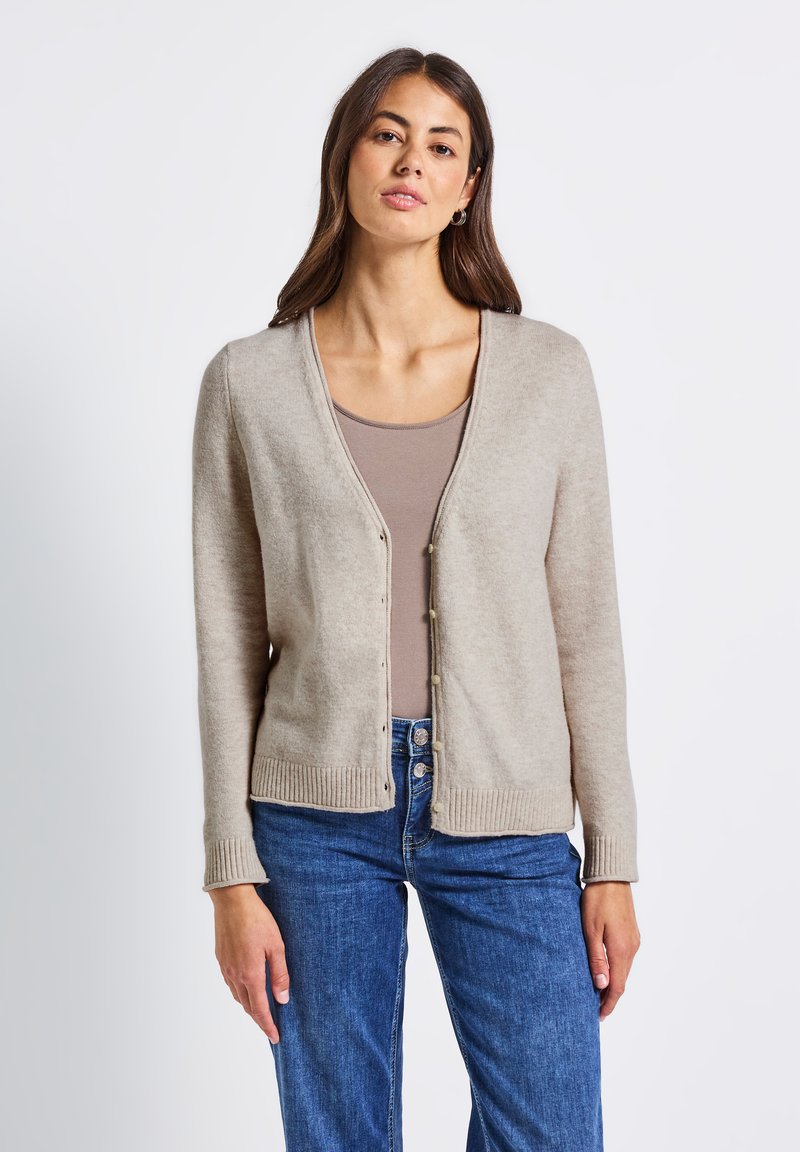 Kvinna iklädd en beige knapplång cardigan över en taupe färgad topp och blå jeans, som står mot en enfärgad vit bakgrund.