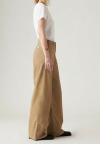 Lehká bílá trička doplněná volnými khaki kalhotami se širokými nohavicemi. Kalhoty mají zadní kapsu a decentní detaily šití. Černá obuv.