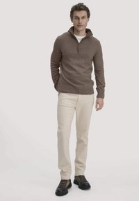 Brauner Strickpullover mit Reißverschlusskragen, kombiniert mit hellbeigen Hosen und dunklen Schuhen. Der Pullover hat eine weiche Textur und eine taillierte Passform.