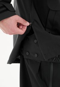 Svart ytterplagg med dragkedjeöppning och metalldrag, samt ett texturerat innerfoder med runda svarta knappar.