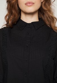 Camisa preta com botões, apresentando pregas verticais e detalhes franzidos nas laterais. Tecido texturizado com colarinho clássico.