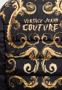 Puuvillane jope, millel on must kangas, kaunistatud julgete, keerlevate kullast ja valgetest mustritest. Esiplaanil on silmapaistev "VERSACE JEANS COUTURE" tekst.
