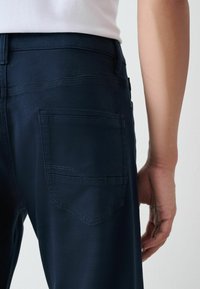 Primo piano del torso inferiore e del braccio di una persona che indossa pantaloni blu scuro con una tasca posteriore e una maglietta bianca infilata dentro.