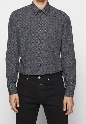 Homme portant une chemise noire à manches longues avec des motifs géométriques blancs, rentrée dans un jean noir, debout devant un fond uni.