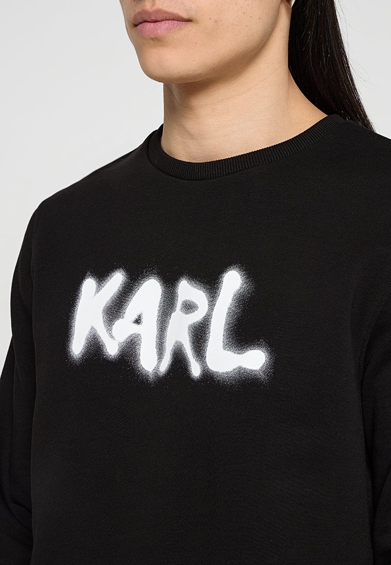 Persona que lleva una sudadera negra de cuello redondo con la palabra "KARL" en un estilo de texto en spray blanco en el pecho, recortada por debajo de la nariz.
