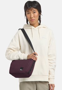 Bordeauxfarbige Schultertasche mit strukturiertem Oberflächendesign, verstellbarem schwarzen Gurt und einem Logopatch. Die Tasche hat eine boxartige Form und ein minimalistisches Design.