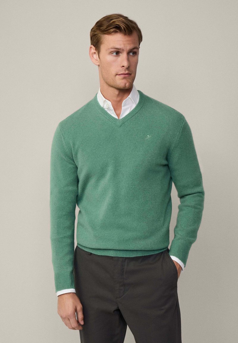 Grön ulltröja med v-ringning och ribbade muddar och nederkant, lagd över en vit skjorta med krage, i kombination med mörka byxor.