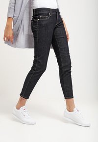 Jeans negros ajustados de denim con herrajes dorados, cinco bolsillos y costuras en contraste. Combinados con zapatillas blancas para un look casual.