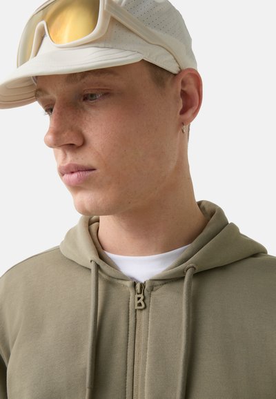 Beige Zip-up-Hoodie mit Kordeln, aus einem strukturierten Stoff, über einem weißen Shirt getragen, vor einem schlichten Hintergrund.