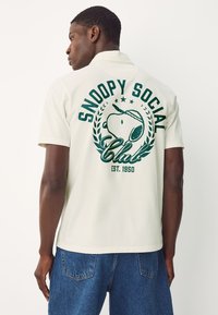 Cremfarbener Poloshirt mit großem grünen Snoopy-Design auf der Rückseite, inklusive dem Text "SNOOPY SOCIAL Club EST. 1950" und Lorbeerakzenten.
